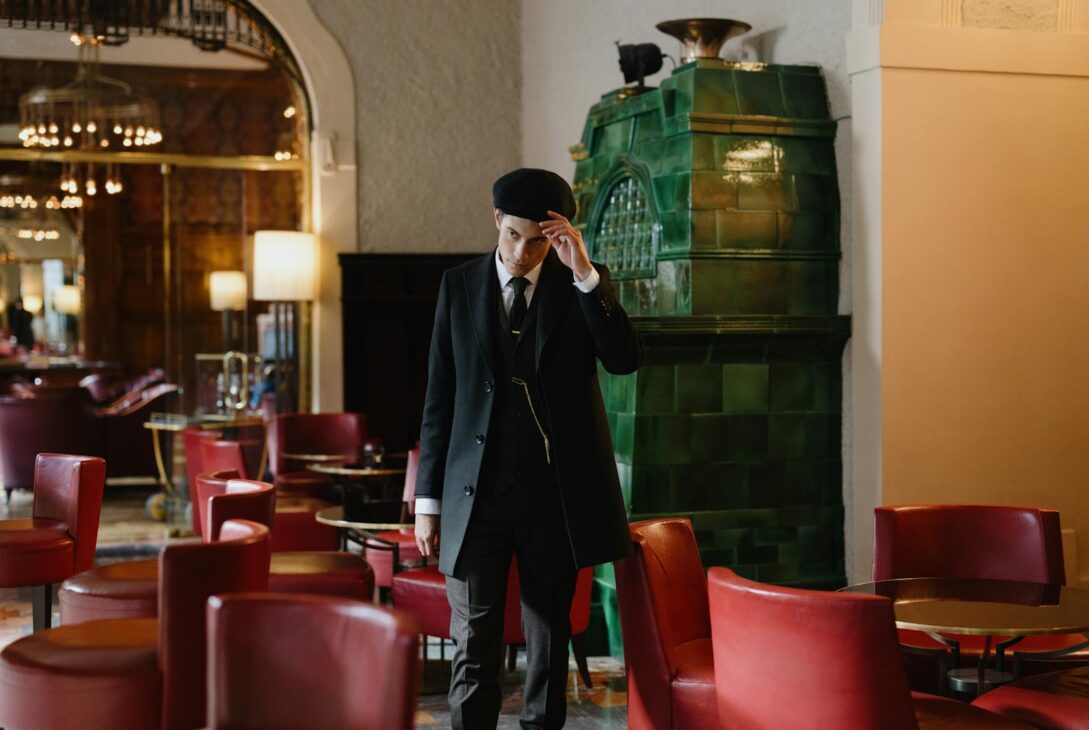 A young man in a black suit and flat cap stands in a stylish vintage bar with red chairs and green stove.