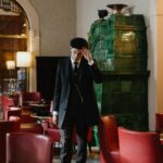 A young man in a black suit and flat cap stands in a stylish vintage bar with red chairs and green stove.