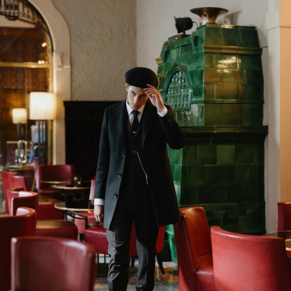 A young man in a black suit and flat cap stands in a stylish vintage bar with red chairs and green stove.