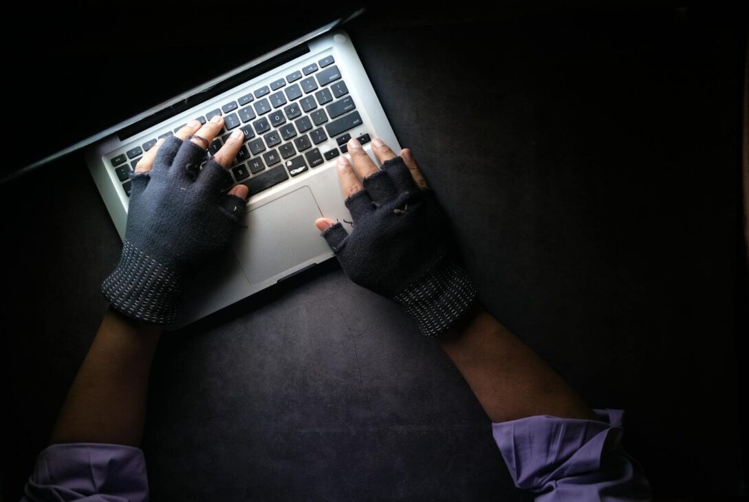 Hacker in fingerless gloves typing on laptop keyboard from above in a dark setting.