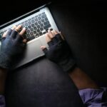 Hacker in fingerless gloves typing on laptop keyboard from above in a dark setting.