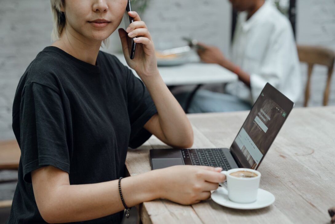 Crop Asian lady in black tee making phone call while sitting at table with coffee and laptop in cafe