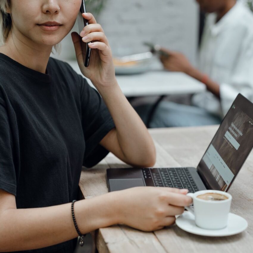 Crop Asian lady in black tee making phone call while sitting at table with coffee and laptop in cafe
